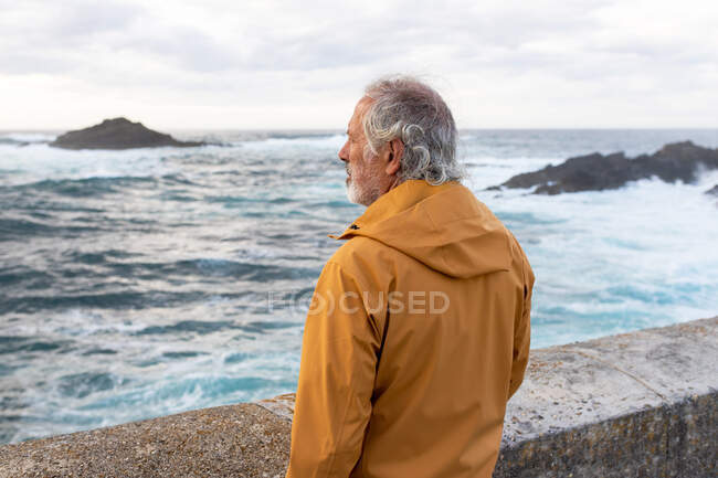 Side view of senior man with gray hair standing on waterfront while admiring stormy sea with rocky formations in coastal area of Spain — Stock Photo