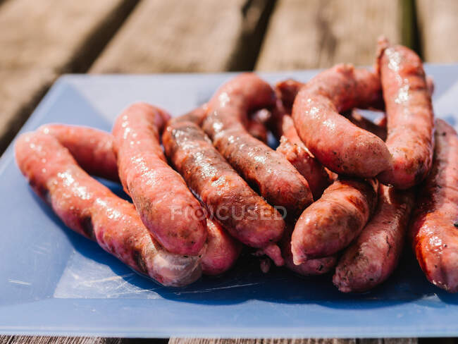 Montón de deliciosas salchichas a la parrilla servidas en la tabla de cortar en la mesa de madera durante la barbacoa en el campo en el soleado día de verano - foto de stock