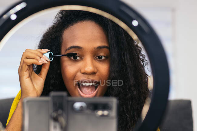 Heureuse femme afro-américaine avec des cheveux bouclés dans des vêtements à la mode appliquant mascara sur les cils avec brosse et enregistrement vlog beauté sur smartphone sur trépied avec anneau lumineux dans la chambre — Photo de stock