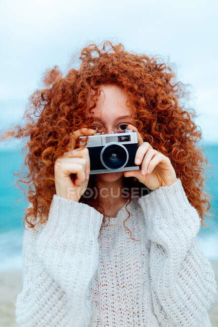 Ginger haired female in knitted sweater looking at camera while taking picture on retro photo camera on coast of sea — Stock Photo