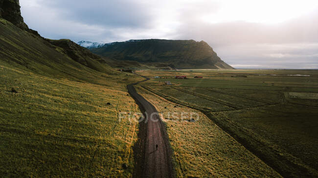 Breathtaking drone view of unrecognizable traveler walking along road near grassy hills during hiking trip in Iceland under cloudy sunset sky — Stock Photo