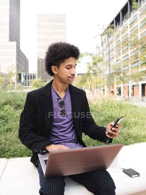 Pensativo jovem trabalhador independente afro-americano cara com cabelos escuros encaracolados em roupas da moda sentado no parque da cidade e trabalhando remotamente no projeto usando laptop e smartphone — Fotografia de Stock