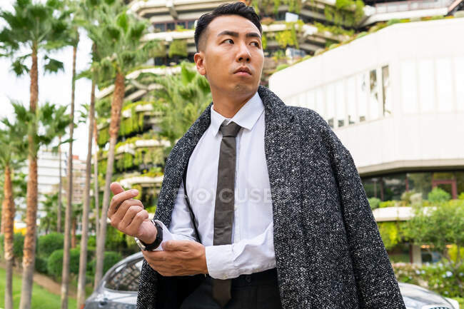 Bem vestido jovem empresário asiático em gravata olhando embora enquanto passeia na estrada contra edifícios modernos na cidade — Fotografia de Stock