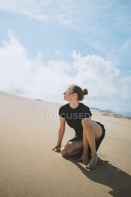 Side view full body of barefoot female leaning on hands while squatting on sandy dune in hot desert — Stock Photo