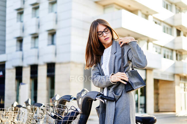 Вид сбоку спокойной женщины в очках и пальто, стоящей у велосипеда на городской улице в солнечный день — стоковое фото