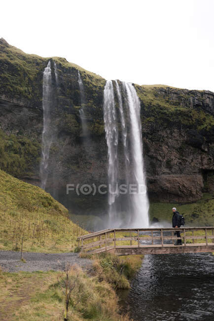 Vue latérale d'un touriste masculin méconnaissable en vêtements chauds et sac à dos debout sur une passerelle en bois et admirant une vue imprenable sur la chute d'eau Seljalandsfoss qui coule à travers une falaise rocheuse dans un étang — Photo de stock