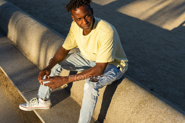 Full body of black man wearing casual t shirt and jeans and white sneakers sitting on concrete border in street at sunlight — Stock Photo