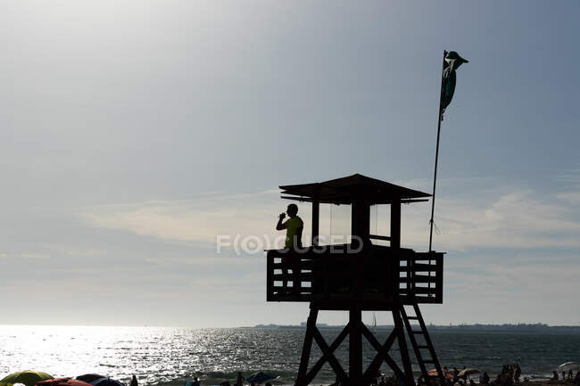 Side view of lifeguard speaking on walkie talkie radio wooden watchtower while overseeing safety at sea against cloudless blue sky — Stock Photo