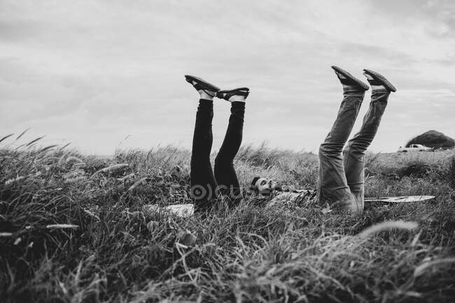 Black and white of side view of cheerful friends in casual clothes lying with raised legs on grassy field in nature in daylight — Stock Photo