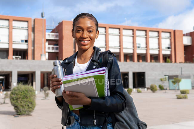 Umtriebige afroamerikanische Studentin mit Thermoskanne und ein paar Schulbüchern blickt in die Kamera, während sie auf der Straße in der Nähe des Universitätsgebäudes steht — Stockfoto