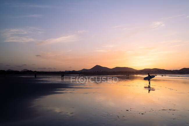 Vista lateral de la silueta de atleta anónima con tabla de surf caminando en el océano ondulado contra la montaña Famara al atardecer en Lanzarote España - foto de stock