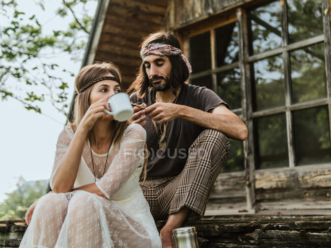Хіпі чоловік дивиться красиву хіпі жінку, що п'є з чашки на дерев'яному ганку — стокове фото