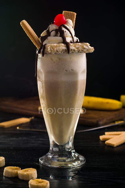 Glass of sweet banana milkshake garnished with whipped cream waffles and cherry with chocolate on top — Stock Photo