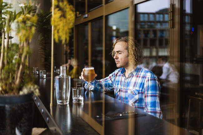 Macho pensativo com cabelo encaracolado em roupas casuais sentado no balcão de madeira perto da janela no bar e bebendo cerveja durante o dia — Fotografia de Stock