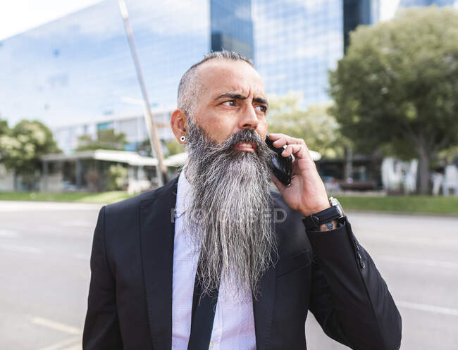 Confident bearded male in classy suit having phone conversation while standing near road on street with modern buildings in city — Stock Photo
