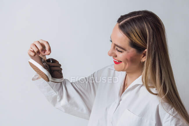 Positive young female in white shirt sitting on bed and preparing baby shoes of childbirth while sitting on bed — Stock Photo