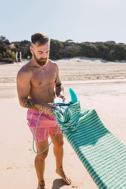 Спортивный человек в купальнике берет защитную сумку с доски для сёрфинга, стоя на песчаном берегу моря в летний день в тропическом курорте — стоковое фото