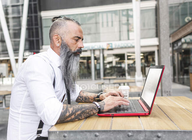 Vista lateral del trabajador masculino hipster reflexivo en netbook de navegación de desgaste formal mientras está sentado en la mesa con café en la terraza de la cafetería - foto de stock