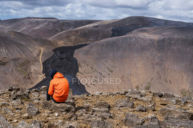 Обратный вид неузнаваемого человека в теплой верхней одежде, сидящего на каменистой земле и любующегося живописными пейзажами действующего вулкана Fagradalsfjall в Исландии при дневном свете — стоковое фото