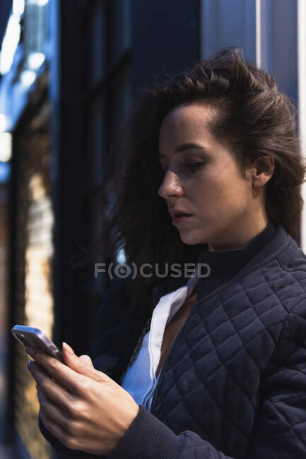 Вид сбоку вдумчивой женщины в теплой одежде с помощью смартфона, стоящей на улице и смотрящей на экран вечером — стоковое фото