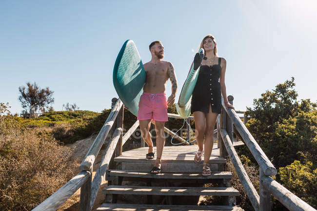 Cuerpo completo de pareja deportiva con tablas de surf paseando por escaleras juntas en camino de madera cerca de plantas verdes antes de entrenar en un resort tropical - foto de stock