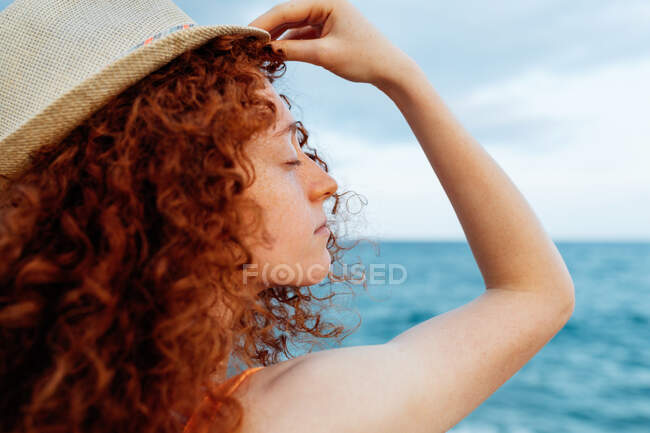 Side view of female with long ginger curls with hat and eyes closed standing on coast of blue sea — Stock Photo
