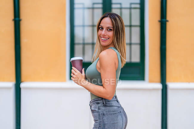 Vista laterale di una donna positiva con tazza di carta di caffè da asporto sorridente e guardando la fotocamera mentre in piedi vicino edificio sulla strada della città — Foto stock