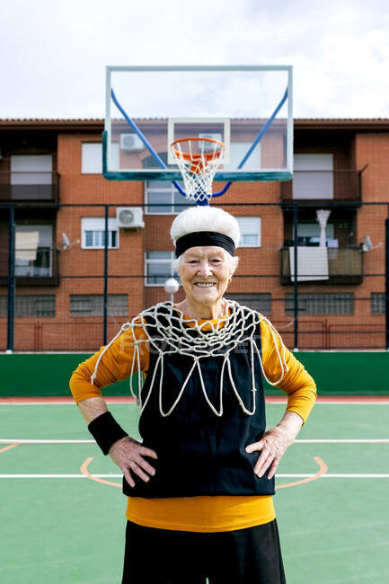 Femme mûre souriante en vêtements de sport et filet blanc regardant la caméra tout en se tenant sur le terrain de sport avec panier de basket-ball pendant l'entraînement — Photo de stock
