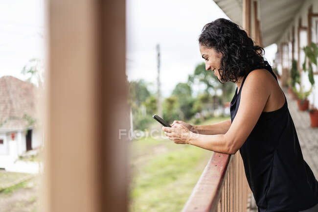 Vue latérale d'une jeune dame ethnique joyeuse aux cheveux noirs bouclés en vêtements décontractés souriant tout en envoyant des messages sur un téléphone portable debout sur la terrasse en bois d'une maison âgée à la campagne — Photo de stock