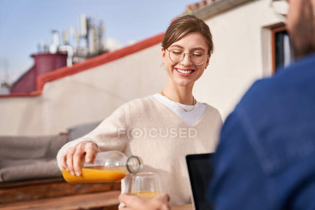 Vue latérale d'une jeune femme positive dans des lunettes versant du jus frais d'une bouteille en verre dans une tasse de colocataire masculin travaillant sur un ordinateur portable sur un balcon — Photo de stock