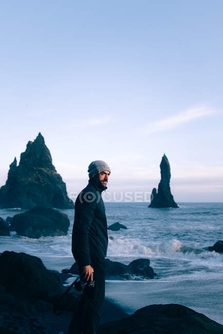 Side view of young bearded ethnic male tourist with photo camera in hand in warm clothes and hat standing on Reynisfjara beach with black sand and admiring Reynisdrangar sea stacks — Stock Photo
