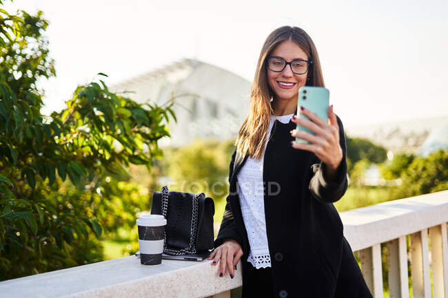 Позитивная женщина в стильном формальном наряде стоящая с ноутбуком и делающая автопортрет на сотовом телефоне рядом с перилами моста — стоковое фото
