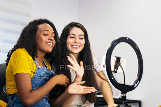 Mujer positiva con el pelo largo y oscuro en ropa casual sentada y mostrando en el alegre blogger afroamericano mientras graba vlog en un teléfono inteligente moderno colocado en el trípode con lámpara de anillo led en casa - foto de stock