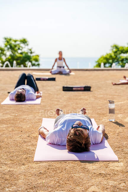Yogalehrer in Aktivkleidung sitzt in Lotusposition, während Menschen bei sonnigem Wetter auf Matten auf dem Boden liegen, während Shavasana im Park stattfindet — Stockfoto