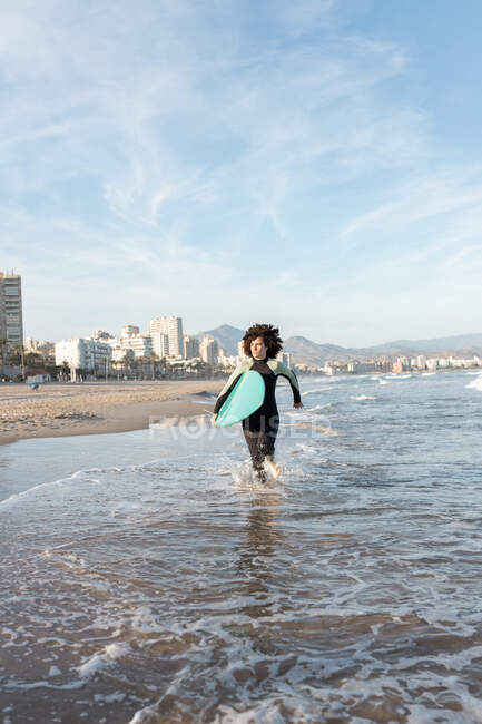 Молодая заботливая серфингистка в гидрокостюме с доской для сёрфинга, бегущая, глядя на берег моря, умытая морем. — стоковое фото