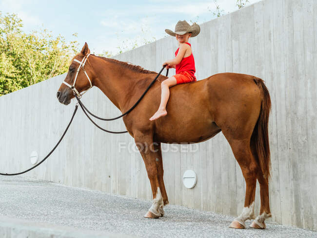 Side view full body of barefoot boy in hat sitting on chestnut horse and holding rein — Stock Photo