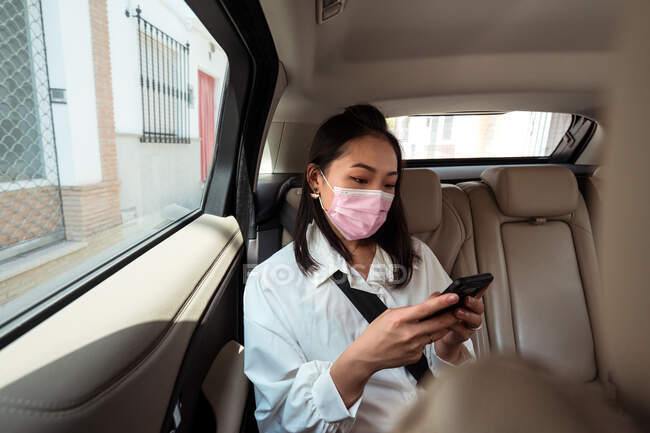 Ethnic female passenger with fastened seat belt using the cell phone while riding in protective mask on backseat in taxi — Stock Photo