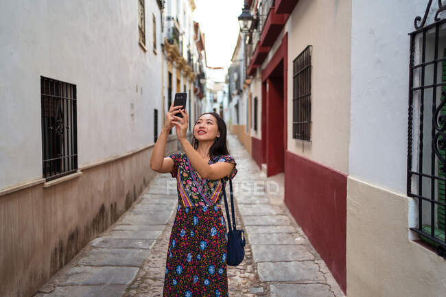 Alegre turista asiática tirando fotos de edifícios envelhecidos enquanto estava em pé na rua estreita pavimentada da cidade de Córdoba, na Espanha — Fotografia de Stock