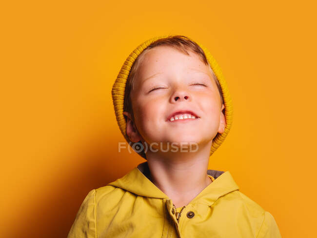 Happy boy in vivid yellow raincoat jacket and beanie hat laughing with eyes closed against yellow background in studio — Stock Photo