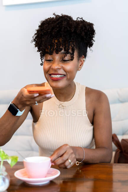 Cheerful young black female millennial with dark Afro hair in stylish clothes taking photo of coffee cup on smartphone while sitting at table in cafe — Stock Photo