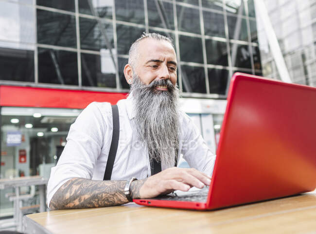 Pensativo hipster trabajador masculino en ropa formal netbook de navegación mientras está sentado en la mesa con café en la terraza de la cafetería - foto de stock