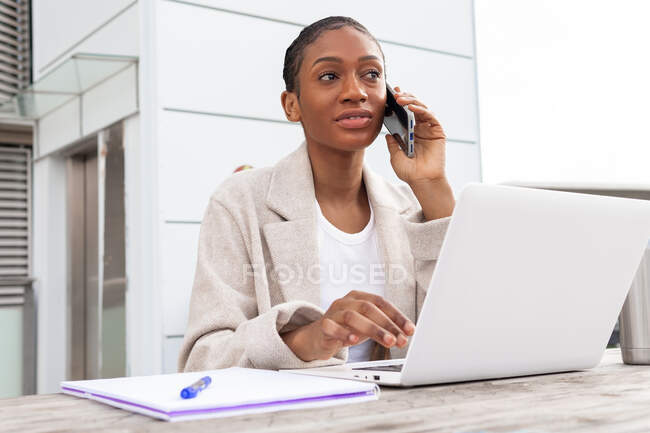 Сфокусована афро-американська жінка-фрілансер озирається, розмовляючи по телефону за столом з нетбуком і записником під час онлайн-роботи на вулиці — стокове фото