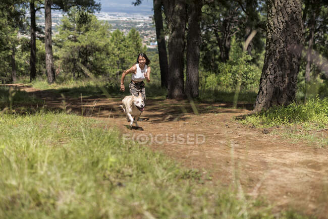 Full body of active female owner running on rural road with loyal dog while training in grove on summer day — Stock Photo
