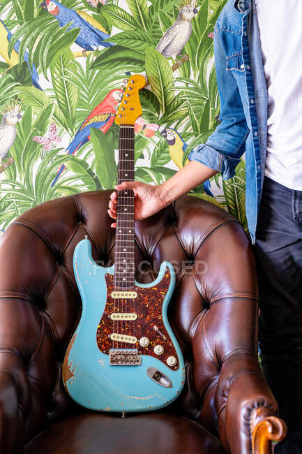 Músico de cultura em roupas casuais colocando guitarra elétrica na poltrona de couro contra a parede colorida vívida — Fotografia de Stock