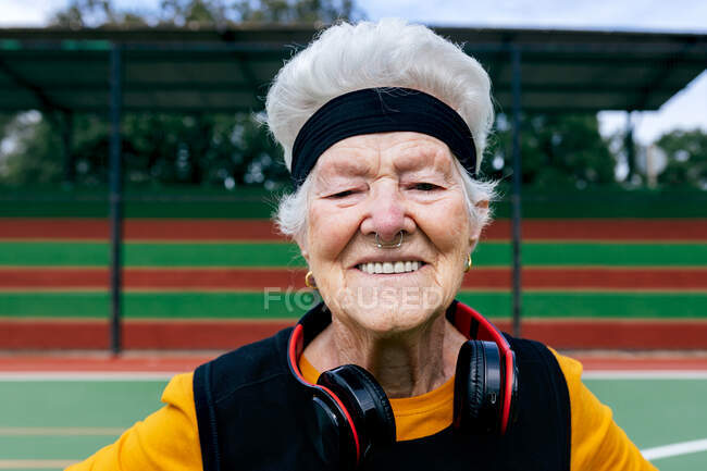 Позитивна зріла жінка з проколотим носом і навушниками в спортивному одязі дивиться на камеру, стоячи на спортивному майданчику під час тренувань — стокове фото