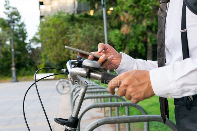 Gestor masculino anónimo de cosecha en ropa formal con bicicleta usando aplicación de uso compartido en el teléfono celular en el pavimento urbano - foto de stock