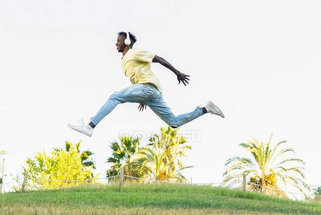 From below full body of young African American male in casual outfit jumping with wide legs apart and hands raised in park — Stock Photo
