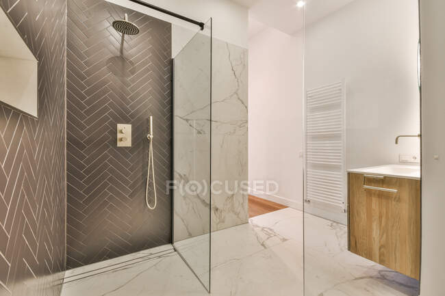Простора скляна душова кабіна та освітлене овальне дзеркало, що звисає на стіні над раковиною у просторій сучасній ванній кімнаті з мармуровою плиткою — стокове фото