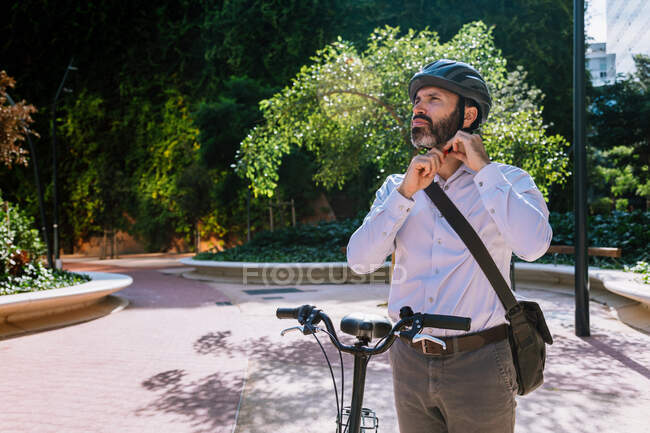 Взрослый бородатый менеджер в формальной одежде, регулирующий шлем для езды на велосипеде в городском парке — стоковое фото