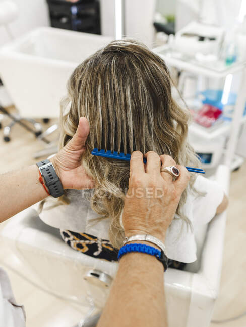 Высокий угол наклона анонимного парикмахера, расчесывающего волосы неузнаваемой клиентки в парикмахерской — стоковое фото